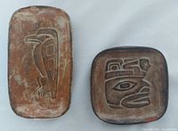 Two rectangular clay plates side by side on white background. Left plate is larger, depicting a raven figure in relief with brown and black patina. Right plate is smaller, depicting a man figure with a 2cm chip on bottom right edge.