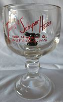 Front view of clear pressed glass goblet with red text 'Tommy's Schuper House 1802 Niagara St. Buffalo N.Y.' and visible black mouse figurine inside near the bottom.
