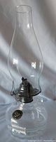 Front view of the Lamplight Farms USA oil lamp with clear tall glass chimney on round clear glass base, metal burner with wick visible