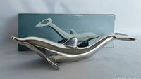 Silverplated dolphin-shaped bottle opener with smooth shiny surface, displayed in front of the light blue box featuring a picture of the opener.