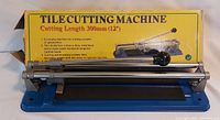 Tile cutting machine in front of its original yellow box showing the product details and specifications.