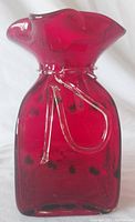 Front and side view of a red blown glass vase with applied clear glass rope detail around neck, dark embedded spots, and with a flared ruffled top edge.