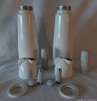 Front view of two white coated metal whipped cream canisters with threaded metal tops, plastic dispenser heads, nozzle tips, caps, and rubber seals laid out in front.