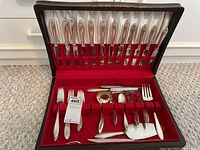 Full view of the open wooden box with 1867 Rogers Bros silver-plated flatware and serving utensils arranged in red velvet slots and with white satin lining in the lid.
