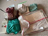 Photo showing folded towels in brown, off-white, and green colors, green bed linens, white plastic bins, and a white embroidered curtain or tablecloth packed in a transparent storage bag.
