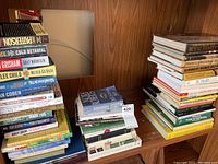 Three stacks of assorted books on wooden shelves, showing both hardback and paperback editions, with various title and topic visible.
