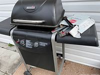 Char-Broil gas BBQ grill with two control knobs and side shelves, some BBQ utensils on the right shelf, against a white siding wall outdoors.
