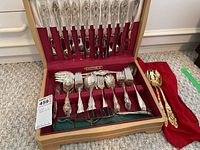 Flatware set stored inside a wooden box with red velvet lining and ivory satin interior lid, showing forks, knives, and spoons arranged neatly.