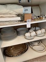 Top shelf with folded table linens and salt and pepper shakers alongside Life@Home dinnerware including stacked plates, cups, and saucers.