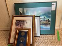 Photo of four framed items stacked against a wall, including a large signed landscape art print and a smaller frame containing three silver coin tokens.