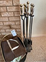 Photo of metal fireplace tools (poker, shovel, broom) with silver-toned handles on black metal stand, next to brass-colored wood holder containing brown material and papers, placed on carpeted floor by brick fireplace.