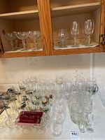 Wide view showing assorted glassware including stemmed wine glasses on shelf and assorted glasses on the counter including beer mugs and water glasses along with wine glass markers in tray.