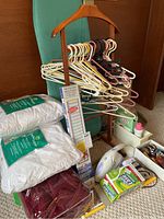 Photo showing an ironing board covered in green fabric, assorted plastic and wooden clothes hangers hanging on a wooden rack, handheld fabric steamer, mini blinds box, and cleaning powders including Dust Ease and Borax with some small miscellaneous items.