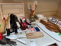 Photo showing various kitchen utensils including rolling pin, can opener, citrus juicer, wooden spoons, masher, tongs, placemats, and ceramic containers.