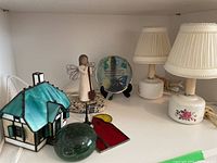 Photo showing two lamps with ceramic bases and fabric shades, a glass house with blue-green roof, wooden angel figurine, glass paperweight with butterfly and text, and stained glass art piece
