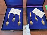 Two open presentation boxes each containing three spoons and a certificate
