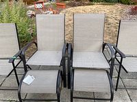 Two patio chairs with beige mesh seats and dark metal frames, each paired with a matching foot rest in front, placed outdoors on stone patio.
