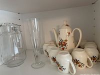 Photo showing ceramic coffee pot decorated with orange and purple floral design along with 11 matching ceramic cups, clear large glass pitcher, tall clear glass vase, and a single tall narrow beer glass.