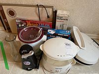 Overall view of lot items on table showing George Foreman grill, Rival crock pot, Black & Decker boxed items, and glass carafe.