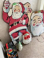 Two large wooden vintage Santa decorations, wooden Santa head, ceramic lighted Christmas tree, and boxed NOMA string lights on carpeted floor against wall.