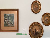 Four framed wall art pieces on a white wall: one larger square wooden framed print depicting a cabin in the forest; three smaller oval gold-toned framed prints with architectural images.