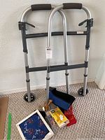 Photo of silver wheeled walker model 6291-5F leaning against wall, with blue Magic Bag foot wrap, wooden massage tools, and other healthcare items placed in front on floor.