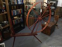 Full side view of the antique wooden spinning wheel showing the wheel, base, and crank handle in a room with other vintage items and books.