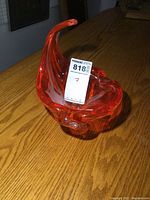 Side angled view of the red-orange Chalet art glass bowl with a curved upward decorative tip on a wooden table.