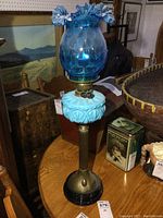 Full view of lamp showing blue ruffled glass chimney and milk glass reservoir on wooden table among other antiques.
