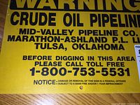 Front view showing yellow sign with black printed warning text about crude oil pipeline and company contact information.