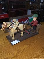 Side view of hand-carved wooden man driving horse figurine on wooden base, showing details of harness and straw bundle.