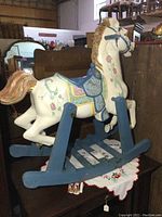 Side view of vintage white wooden rocking horse with floral decoration and blue saddle on blue rockers.