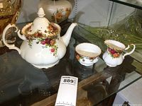 Teapot, sugar bowl, and creamer displayed together on glass shelf, showing floral pattern and gold accents.
