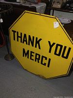 Front view of yellow octagonal metal sign with "THANK YOU MERCI" in black.