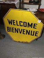 Front view of the vintage yellow metal octagon sign with black 'WELCOME BIENVENUE' lettering showing wear and rust at the bottom edge.