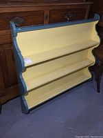 Front view of the three-tier painted wood display shelf showing yellow shelves and blue-green trim, with minor wear visible.