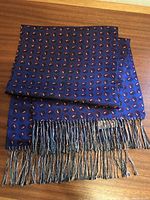 Folded deep royal blue silk scarf with burgundy and cream paisley design, showing hand-tied multi-colored silk fringe ends, placed on wooden surface.