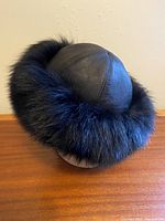Side angled view of black leather hat with fluffy long hair mink fur trim around the edges, showing the smooth leather upper and thick fur border.