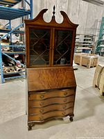 Full front view of the antique serpentine drop down secretary desk showing the arched pediment top, glass display case with geometric wooden design, drop front writing surface, and four drawers with original batwing handles.