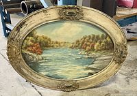 Full view of the oval framed waterscape oil painting showing the river, trees, and ornate carved frame with aged patina and wear.