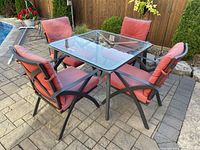 Photo showing three wrought iron chairs with red cushions arranged around a square wrought iron table with a glass top outdoors on stone patio.