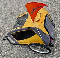 Side angle view showing the yellow and gray fabric construction, mesh windows, clear top window, zippered pet door/storage pouch, black coupler/tow bar, and large spoked wheels with rubber tires. Orange Schwinn flag attached at the rear.