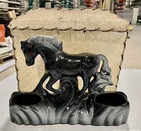 Front view of black glazed pottery horse lamp with two compartments and parchment paper shade behind it.