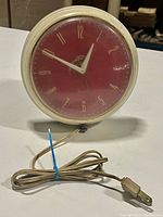 Front view of vintage Westclox wall clock with plugged-in power cord showing red face plate and clear cover.