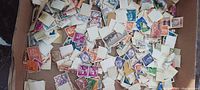 Hundreds of loose assorted postage stamps in a flat container, visible variety of colors, postmark presence, and perforated edges