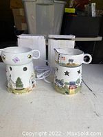 Two ceramic electric simmer pots with different decorative painted outdoor scenes. Each has star-shaped cutouts and power cords visible with white cardboard boxes behind.