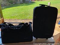 Two black Freedom Bag 2000 pieces: a duffel bag on the left and a wheeled suitcase on the right, placed on a table with grass visible outdoors in the background.