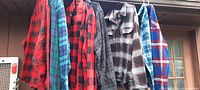 Seven men's long sleeve shirts hanging on a rack outdoors, various plaid patterns, colors include red, black, blue, grey