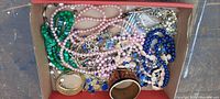 Photo showing assorted necklaces, bracelets, and pins including beaded necklaces in green, pink, blue and mixed colors, a wide wooden/enamel floral bangle, and tan cuff bracelet inside a box.
