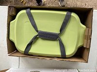 Top-down view of light green baking dish held with grey straps inside cardboard box.